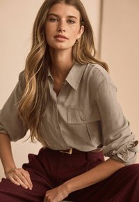 Light gray button-up shirt with rolled sleeves and two front pockets, paired with burgundy striped high-waisted trousers. Simple, textured fabric.