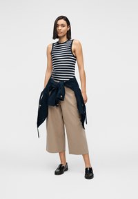 Ριγέ αμάνικη μπλούζα σε ναυτικό μπλε και λευκό, συνδυασμένη με φαρδιές μπεζ παντελόνες και ναυτικό μπλέ σακάκι δεμένο γύρω από τη μέση. Μαύρα loafers ολοκληρώνουν το σύνολο.