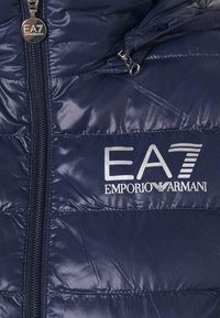 Chaqueta acolchada azul marino con cierre de cremallera, textura brillante acolchada y un parche con el logotipo "EA7 Emporio Armani" en la parte delantera.