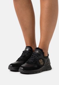 Black athletic shoes with a combination of smooth and textured materials, featuring a faux fur collar, logo accents, and a lace-up design.