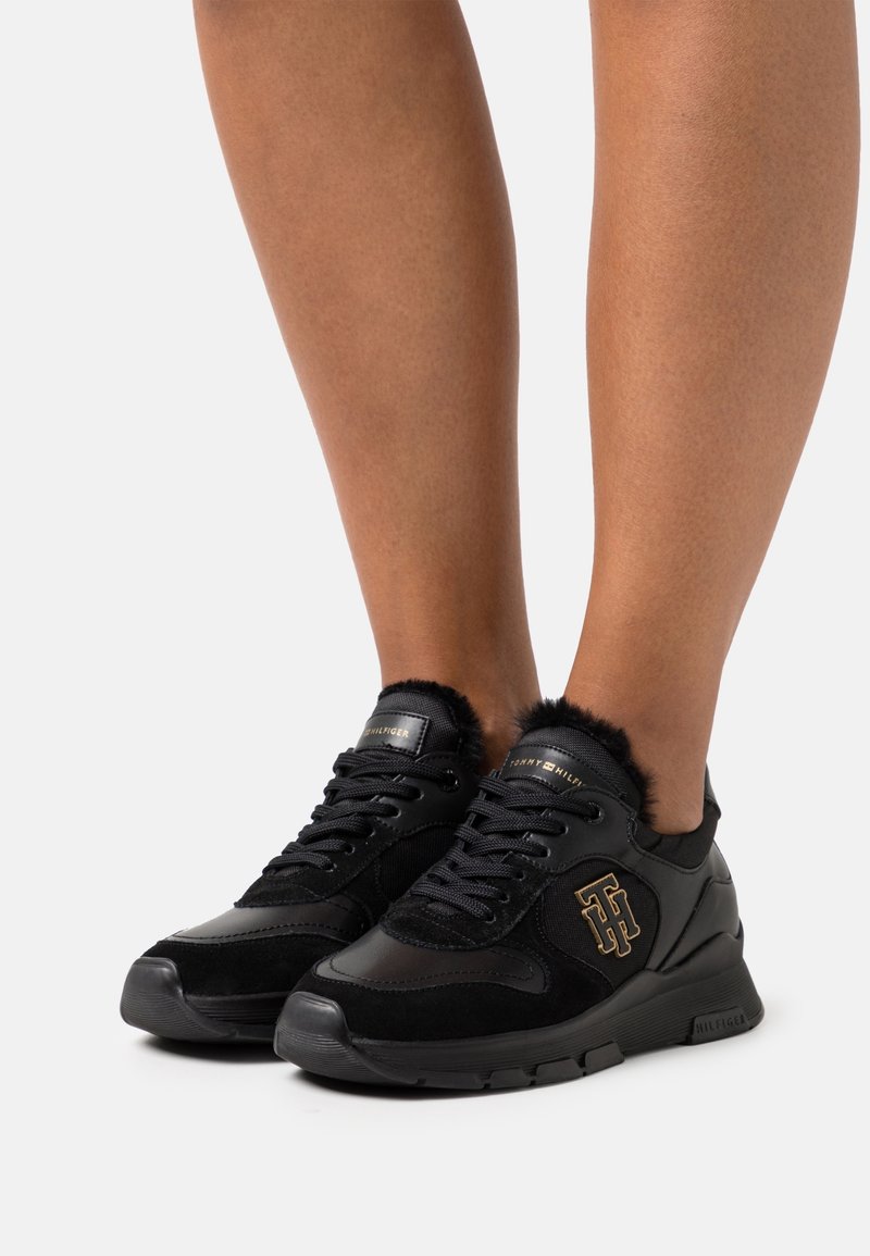 Black athletic shoes with a combination of smooth and textured materials, featuring a faux fur collar, logo accents, and a lace-up design.