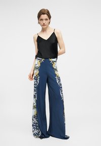 Γυναίκα που φοράει μαύρο αμάνικο τοπ και ψηλόμεσα φαρδιά μπλε παντελόνια με χρυσά και λευκά διακοσμητικά μοτίβα, στέκεται μπροστά σε λευκό φόντο.