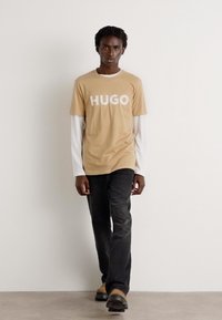 Šviesiai ruda marškinė su balta "HUGO" logotipu, uždėta ant baltų ilgomis rankovėmis marškinėlių. Pora su juoda džinsais ir tamsiais bateliais, stovintis prieš neutralų foną.