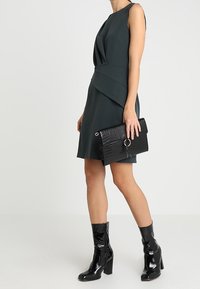 Ciemnozielona sukienka bez rękawów z drapowanym wzorem, zestawiona z czarną kopertówką z tłoczeniem imitującym skórę krokodyla oraz lakierowanymi botkami za kostkę.