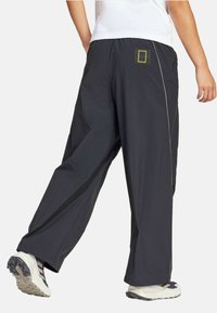Crne hlače širokih nogavica s glatkom teksturom, šivanim detaljima i brendiranom etiketom na stražnjoj strani. Uparene s svijetlim cipelama.