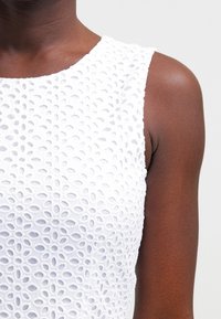 Biały top bez rękawów z teksturowanym wzorem owalnych wycięć, gładką tkaniną i okrągłym dekoltem, ukazujący dopasowany krój.