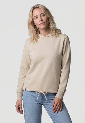 Blondes Frau trägt einen beigen Hoodie mit Kordelzug und hellblaue Jeans, steht vor einem schlichten hellen Hintergrund.