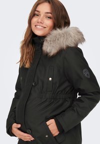 Juoda parka su kailine kapišonu, mygtukų uždarymu ir surinkta juosmeniu. Yra lopinėlio logotipas ant rankovės ir šoninės kišenės.