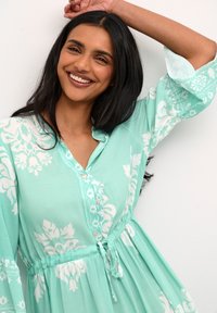 Svetlo zelena obleka z belimi cvetličnimi vzorci, gumbi in pasom na vrvico. Razširjene rokave dodajajo volumen oblikovanju.