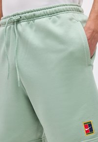 Ανοιχτό πράσινο φόρμας με κορδόνι στη μέση, πλευρικές τσέπες και ένα μικρό πολύχρωμο μπατζ Nike κοντά στο τελείωμα στο αριστερό πόδι.