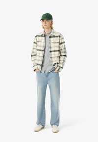 Un giovane indossa un abbigliamento casual: giacca a quadri, camicia a righe, jeans chiari e berretto verde, sorridendo davanti a uno sfondo bianco semplice.
