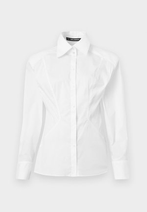 Chemise pour femme blanche à manches longues, ajustée, avec col pointu, fermeture à boutons sur le devant et détails discrets de coutures sur les côtés.