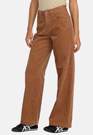 Femme portant un pantalon marron en velours côtelé à taille haute et jambes larges, associé à des baskets noires et blanches, debout devant un fond uni.