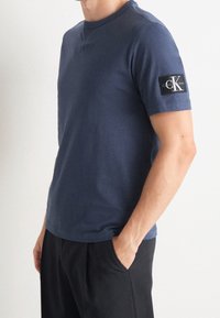 Mies, który ma na sobie granatową koszulkę krótko rękawową Calvin Klein oraz czarne spodnie, trzyma rękę w kieszeni i stoi na jednolitym tle.