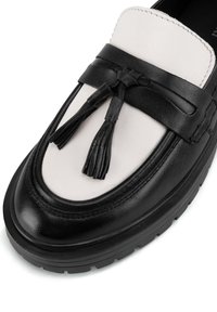 Černobílé kožené mokasíny s zaoblenou špičkou, ozdobnými černými třásněmi a texturovanou gumovou podrážkou pro lepší přilnavost.
