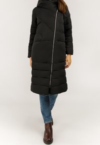Abrigo puffer negro con cuello alto, patrón acolchado horizontal, cierre frontal, dos bolsillos laterales y mangas largas. Combinado con jeans y botas.