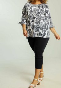 Blouse à fleurs noire et blanche avec manches trois-quarts et encolure froncée, associée à un pantalon noir coupé au-dessus de la cheville et des talons blancs à lanières.