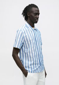 Īsroku krekls ar zilām un baltām vertikālām svītrām, ar apkakli, kabatu un fakturētu audumu, kombinēts ar gaišas krāsas biksēm.