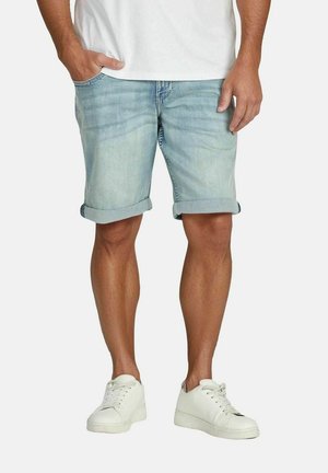 Mann trägt hellblaue, hochgekrempelte Jeansshorts, weiße Turnschuhe und ein weißes T-Shirt, steht mit einer Hand in der Tasche.