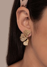 Oreja con un pendiente metálico dorado en forma de dos hojas de abanico superpuestas sobre un fondo neutro.