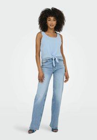 Ljusblå ärmlös topp med en framknutningsdetalj, ihop med ljus tvättade utsvängda jeans och klackar med öppen tå. Slät tygtextur.