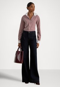 Γυναίκα που φοράει κόκκινο και άσπρο ριγέ πουκάμισο με φαρδιά γιακά, σκούρα φαρδιά τζιν, καφέ τακούνια, κρατώντας μια υφασμάτινη σκουρόχρωμη κόκκινη τσάντα.