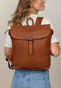 Brauner Leder-Rucksack mit Klappenverschluss, vorderem Riemen-Detail und verstellbaren Schulterriemen. Glatte Textur, minimalistisches Design.