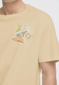 Light beige cotton t-shirt featuring a printed design with a newspaper, lemon, croissant, and coffee cup. Short sleeves, round neckline.