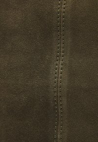 Tissu en daim vert olive foncé avec une texture lisse. Présente une couture apparente avec des points contrastants le long du centre.