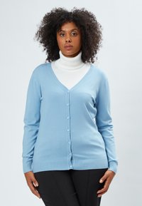 Helles blaues Cardigan aus weichem Stoff, mit V-Ausschnitt, Knopfverschluss, gerippten Bündchen und Saum. Wird über einem weißen Rollkragenpullover getragen.