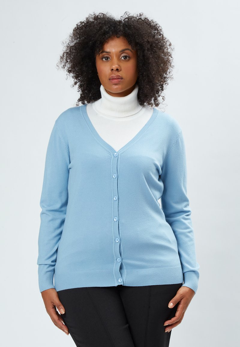 Helles blaues Cardigan aus weichem Stoff, mit V-Ausschnitt, Knopfverschluss, gerippten Bündchen und Saum. Wird über einem weißen Rollkragenpullover getragen.