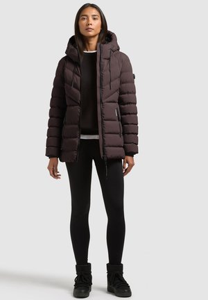 Une femme porte une doudoune à capuche marron foncé, un legging noir et des bottes sur un fond clair et uni.