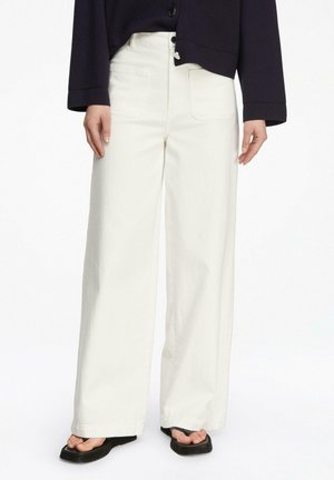 Osoba ubrana w białe, szerokie spodnie z wysokim stanem, w zestawieniu z czarną, zapinaną na guziki kurtką oraz czarnymi sandałami z kwadratowym noskiem.