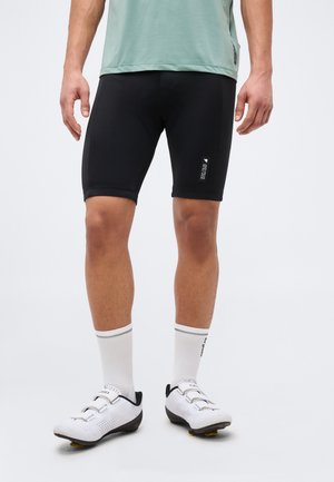 Férfi fekete kerékpáros rövidnadrágban, fehér zokniban és fehér, tüskés kerékpáros cipőben, egy egyszínű világos háttér előtt áll.