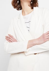 Bílý blejzr s dlouhými rukávy, výstřihem s vykrojením a dvěma předními kapsami. Hladká látka a uvolněný střih, vrstvený přes proužkované tričko.