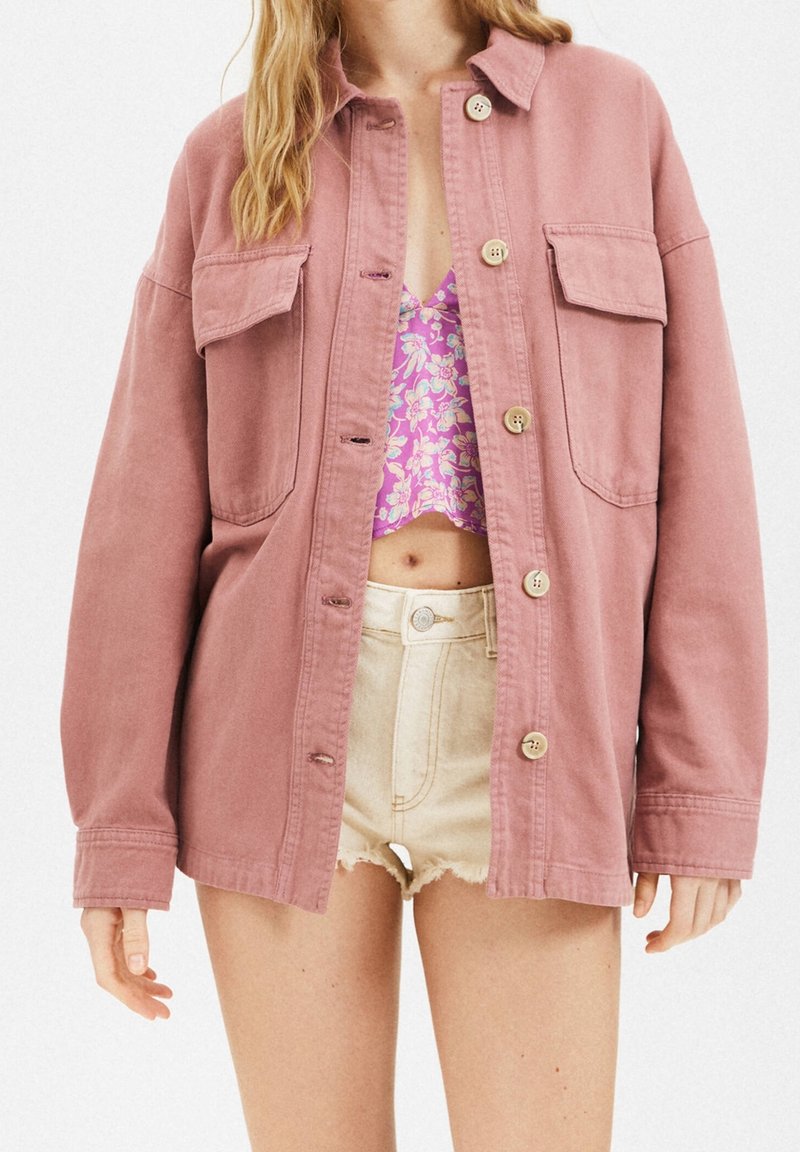 Różowa, oversize'owa kurtka wykonana z bawełny, z dwoma przednimi kieszeniami, zapięciem na guziki i luźnym krojem. Noszona na kwiecistym crop topie.