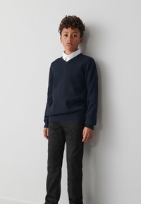 Tmavomodrý sveter zo zmesi vlny s výstrihom do V, nosený cez bielu košeľu s golierom, v kombinácii s čiernymi nohavicami. Jednoduchý a klasický dizajn.