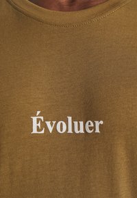 Tissu marron avec le mot brodé en blanc "Évoluer" centré sous un col rond.