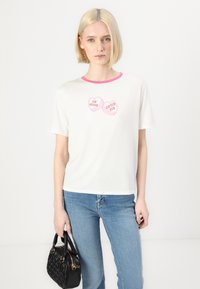 T-shirt de algodão branca com mangas curtas, decote redondo rosa e dois gráficos em forma de coração com texto. Combinado com jeans azul claro e mala preta.