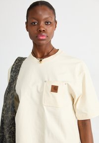 Crème katoenen T-shirt met korte mouwen, een borstzak met een bruine logopatch en een gestructureerde grijze schoudertas.