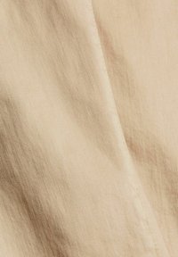 Beige tkanina s texturovaným povrchem, s diagonálními vzory tkaní a jemným leskem; viditelné švy podél okrajů.