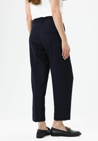 Donkerblauwe, hooggetailleerde broek met een losse pasvorm, voorzien van een rekbare tailleband, twee achterzakken en taps toelopende pijpen. Gecombineerd met zwarte loafers.