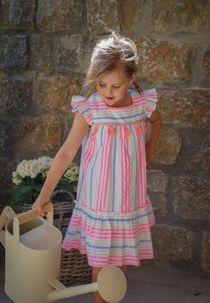 Gestreiftes Kleid mit Rüschenärmeln in Pastellrosa, Blau und Gelb. Hält eine hellfarbene Metallgießkanne mit rundem Ausguss.