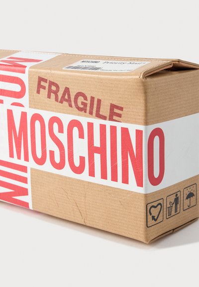 Boîte d'expédition en carton emballée avec du ruban blanc indiquant "MOSCHINO" et "FRAGILE" en rouge, affichant des icônes de manutention et une étiquette de courrier prioritaire attachée.