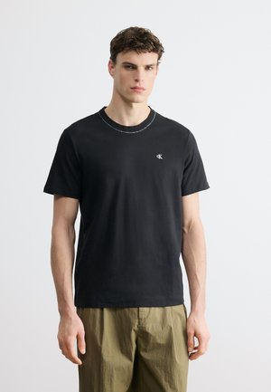 Mężczyzna ubrany w gładką czarną koszulkę z krótkim rękawem z małym białym logo na piersi i oliwkowe zielone spodnie, stojący na białym tle.