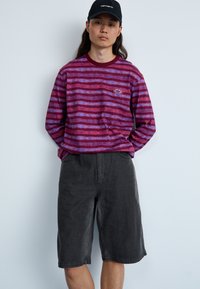 Camisa de manga larga a rayas moradas con cuello y logotipo en contraste color burdeos, combinada con pantalones cortos de mezclilla grises. Diseño simple y casual.