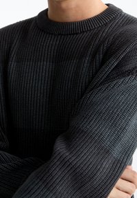 Personne portant un pull en maille côtelée gris foncé, les bras croisés, montrant la texture détaillée du tissu et les manches longues.