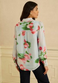 Bluse med blomsterprint, der har en lyseblå baggrund med mønstre af pink og grønne blomster, lange ærmer og en manchet med knap. Glat stof.