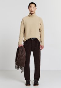 Maglione girocollo beige texture, pantaloni in velluto a coste marrone e scarpe in pelle marrone scuro, mentre tiene una sciarpa marrone con frange.