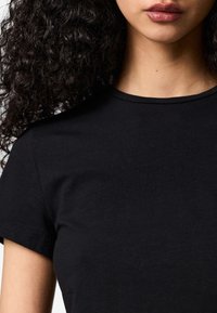 Persona con capelli ricci e carnagione media che indossa una t-shirt nera a maniche corte e scollo rotondo, mostrata dal mento alla parte superiore del torso.